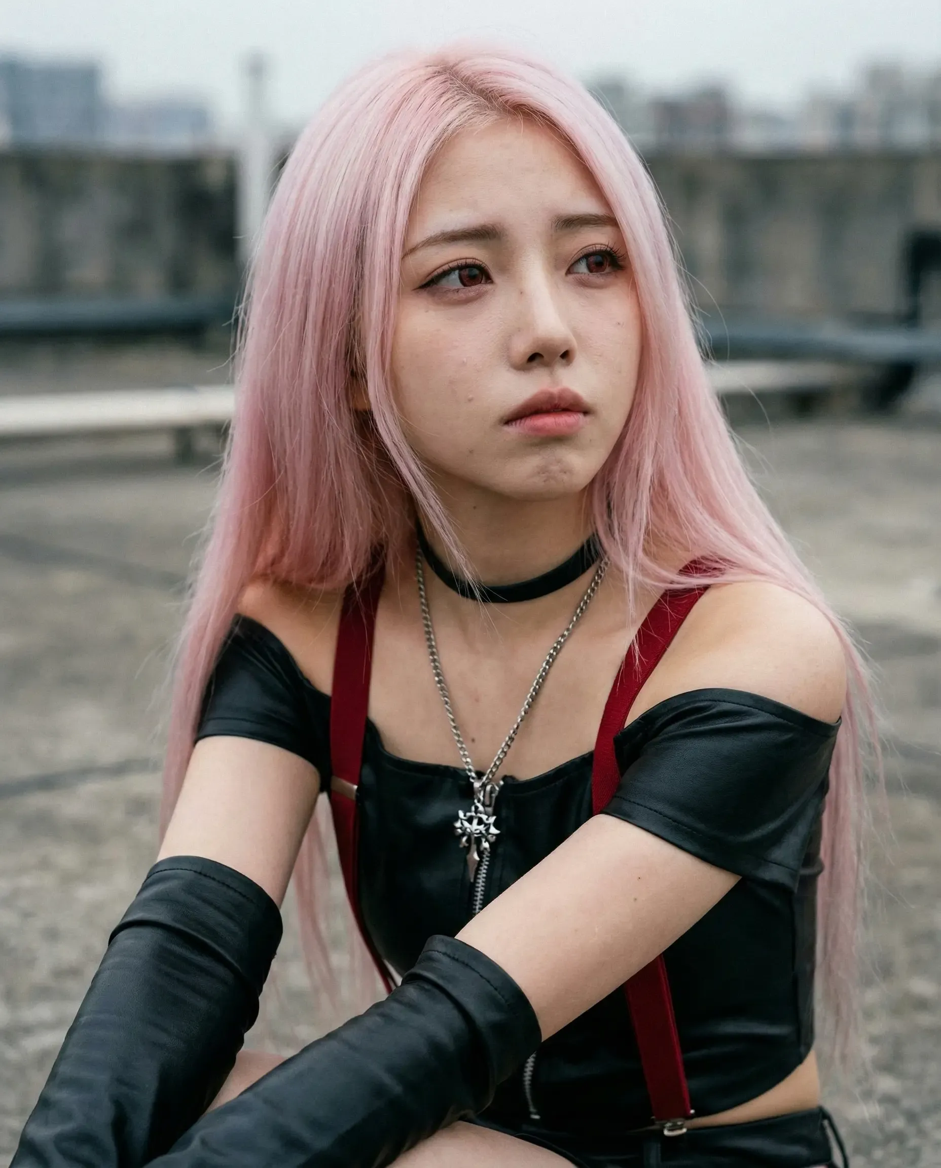 ピンクの長髪に黒い衣装と赤いサスペンダーを着用し、屋外で遠くを見つめる女性