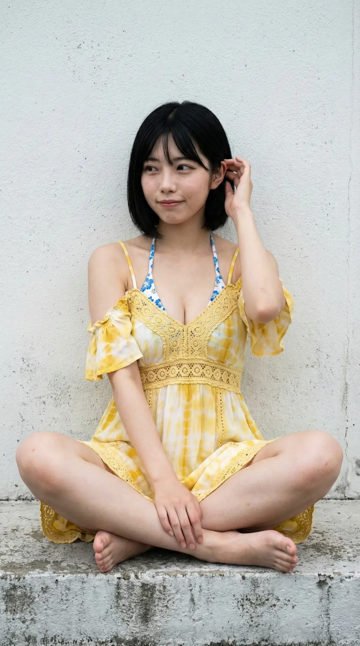 白い壁の前で黄色いタイダイ柄のワンピースを着て、あぐらをかいて座る黒髪の女性。
