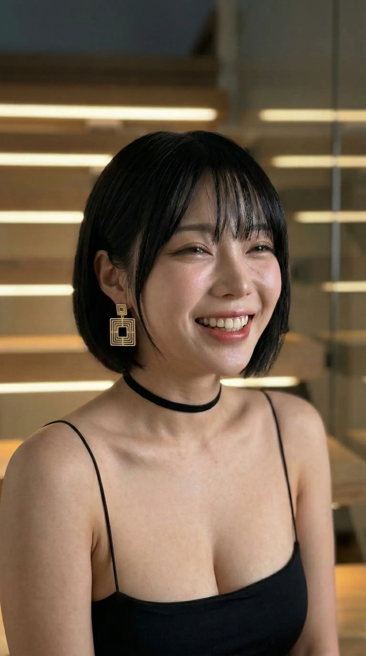 黒髪ボブで笑顔の女性。黒いキャミソール、チョーカー、四角い金のイヤリングを着用。