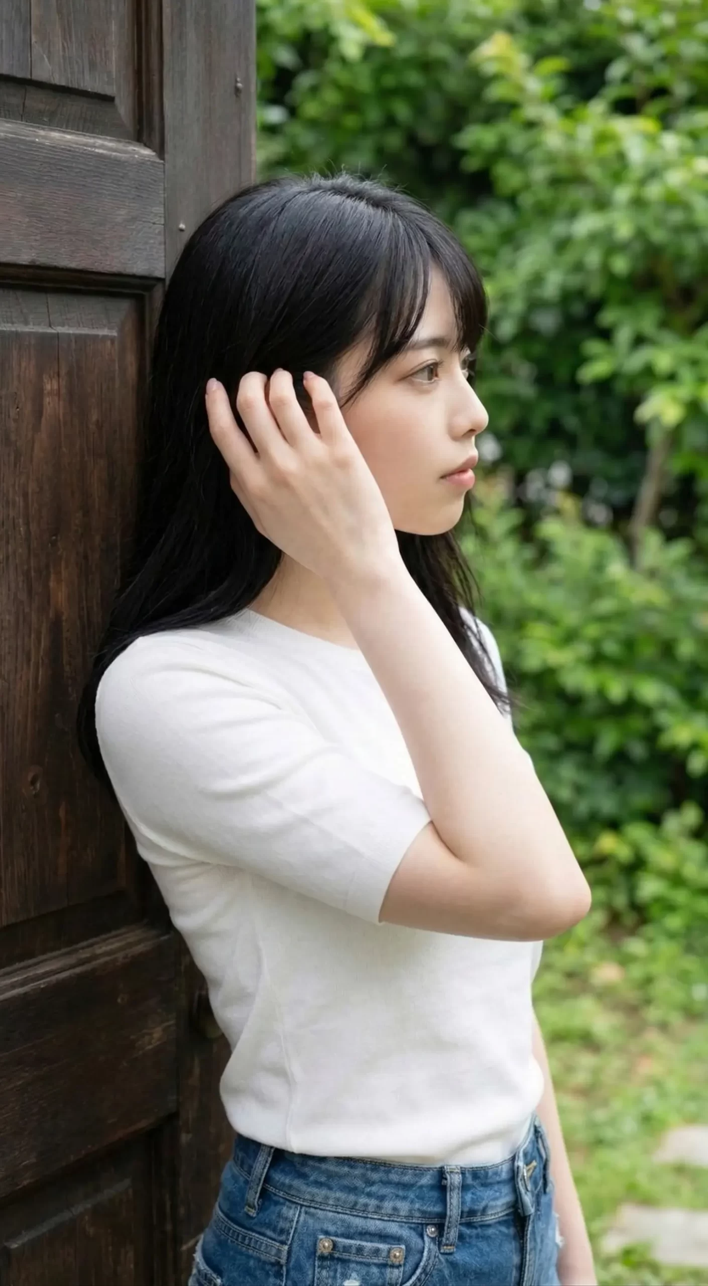 白いトップスを着た黒髪の女性が、木の扉の横で髪に手を添えながら横を向く姿。