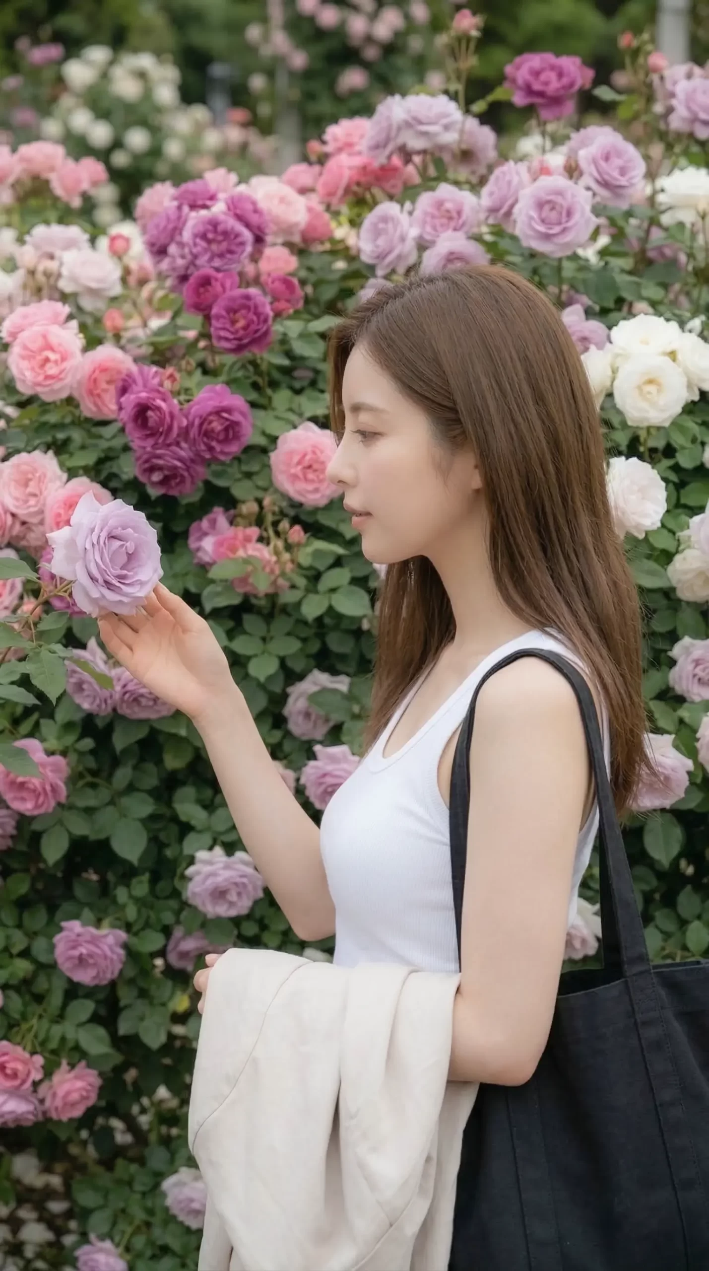 満開のバラ園で、薄紫色のバラに手を添えて見つめるロングヘアの女性。