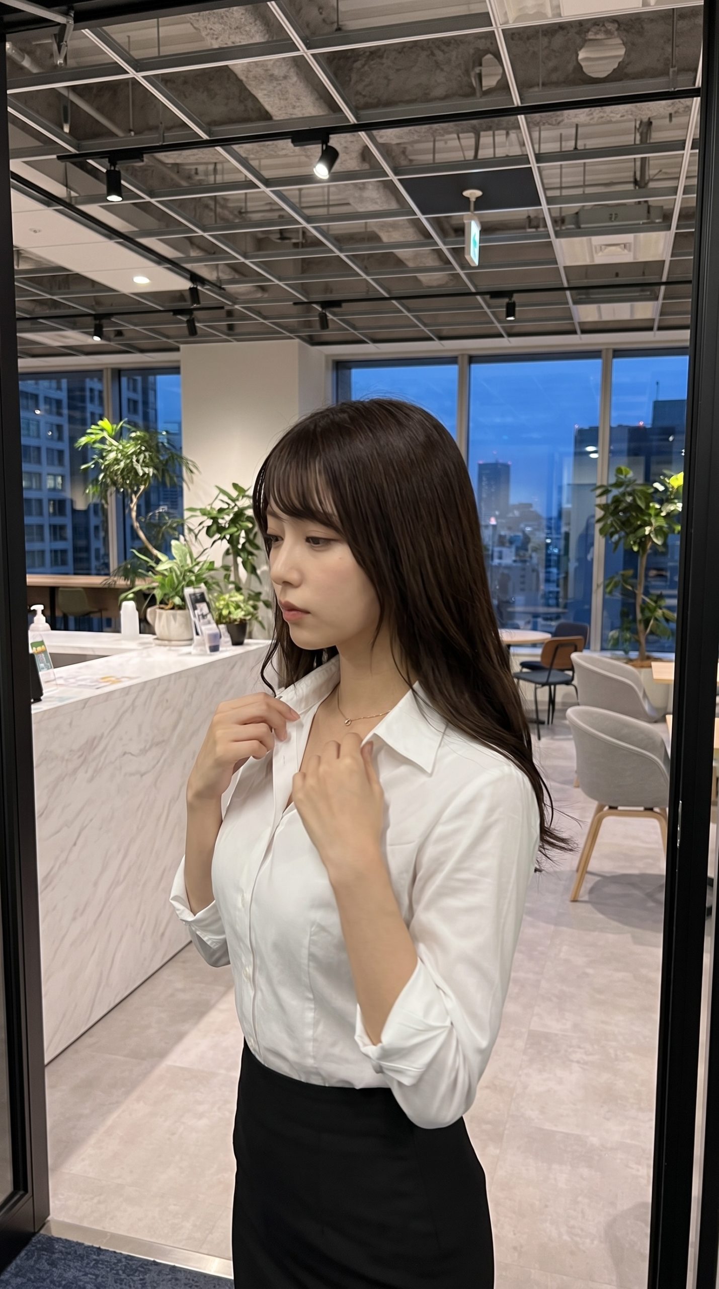 オフィスの窓際で白いシャツの襟元を整えるロングヘアの女性